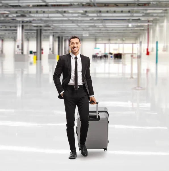 スーツケース付きの空の空港ホールを歩く黒いスーツケースの笑顔の若い男の完全な長さの肖像画 — ストック写真