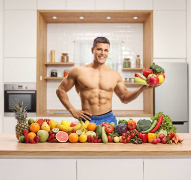 Genç kaslı adam, modern bir mutfağın içinde tepsi ve sebze yığınıyla üstsüz poz veriyor.