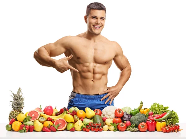 健美运动员摆姿势 拿着一堆水果和蔬菜 指着被白色背景隔开的腹肌 — 图库照片