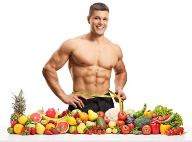 Belini ölçen ve beyaz arka planda izole edilmiş sağlıklı meyve ve sebze yığınıyla poz veren kaslı bir adam.