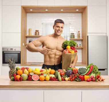 Elinde kese kağıdıyla mutfakta üstsüz bir adam ve tezgahın üzerinde meyve ve sebze yığını. 