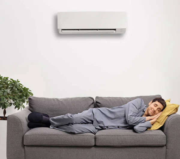 Man in pajamas sleeping on a sofa under an air conditioning unit isolated on white background