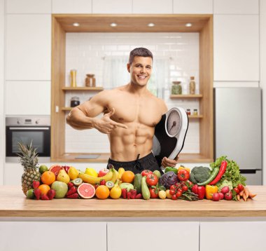 Ahşap tezgahta çeşitli meyve ve sebzeler ve mutfakta örgü ören terazili üstsüz bir adam.