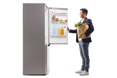 Bakkal çantalı bir adam beyaz arka planda izole edilmiş buzdolabı açıyor.