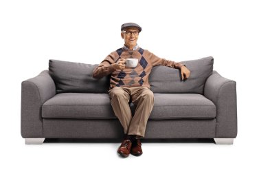 Yaşlı bir adam beyaz arka planda bir fincan çayla kanepede oturuyor.