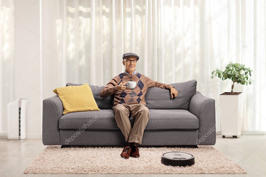 Robotic vacuum cleaner cleaning the floor and an elderly man drinking tea on a sofa at home