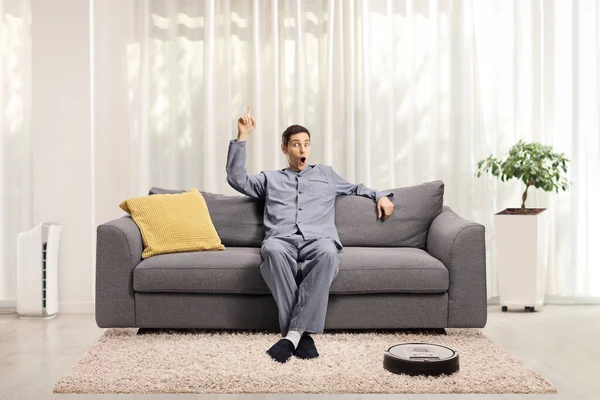 Young Excited Man Pajamas Seated Sofa Home Pointing Robotic Vacuum — Stock Photo, Image