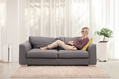 Dizüstü bilgisayarda çalışan ve evdeki kanepede yatan genç bir kadının profil fotoğrafı.