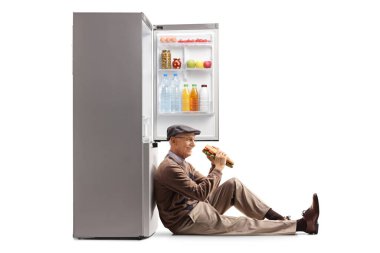 Yaşlı adam sandviç yiyor, yerde oturuyor ve beyaz arka planda izole edilmiş açık bir buzdolabına yaslanıyor.