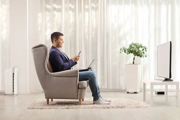 リビングルームに座っているノートパソコンと携帯電話を持つアームチェアの男 — ストック写真