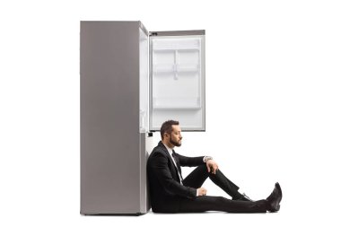 Hayal kırıklığına uğramış işadamı yerde oturuyor ve beyaz arka planda izole edilmiş boş bir buzdolabına yaslanıyor.