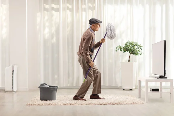 一名老年男子在家里的电视机前 拿着清洁拖把 一边唱歌一边跳舞 — 图库照片