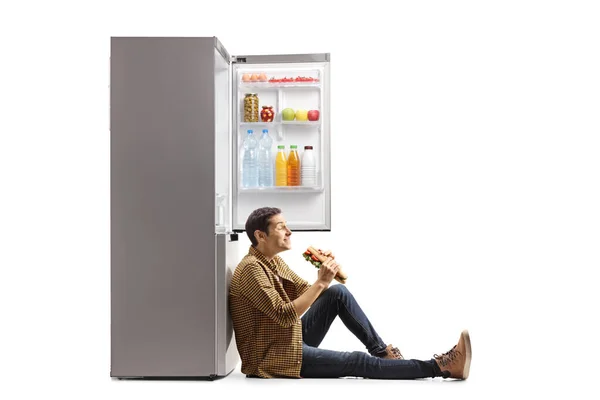Hungry young man with a baguette sandwich leaning on  a fridge isolated on white background