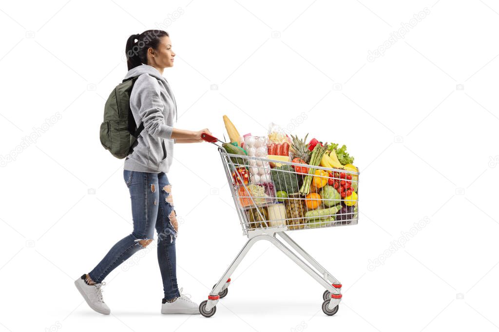 Full length profile shot of a female student walking and pushing a full shopping cart isolated on white background