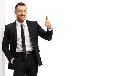 Siyah takım elbiseli bir iş adamı duvara yaslanıyor ve beyaz arka planda izole edilmiş baş parmaklarını gösteriyor.