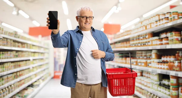 Homem Maduro Supermercado Mostrando Telefone Celular Carregando Uma Cesta Compras — Fotografia de Stock