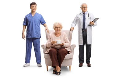 Doktor ve bir sağlık görevlisi, beyaz arka planda izole edilmiş bir koltukta bir fincan çayla yaşlı bir kadının yanında duruyor.