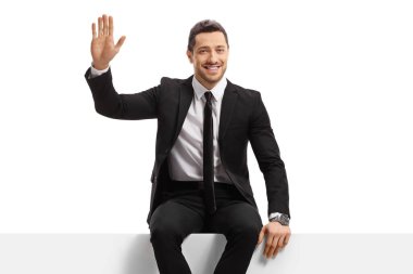 Gülümseyen işadamı bir panelde oturuyor ve beyaz arka planda izole edilmiş kameraya el sallıyor.