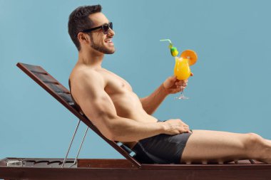 Mavi arka planda izole edilmiş güneşli bir yatakta kokteyl içen genç adam.