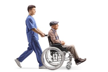 Beyaz arka planda, tekerlekli sandalyedeki yaşlı bir adamı iten erkek hemşirenin tam boy profil fotoğrafı.