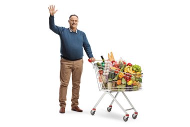 Elinde alışveriş arabası ve beyaz arka planda izole edilmiş kameraya el sallayan yetişkin bir adamın tam boy portresi.