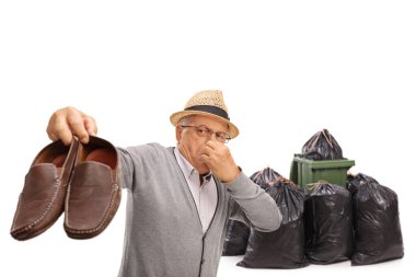 Elderly man holding a pair of stinky shoes near a waste bin and bags isolated on white background clipart