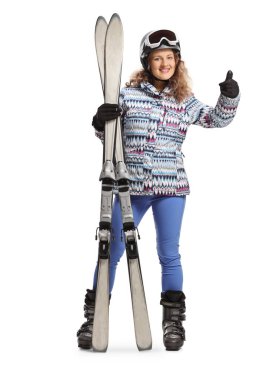 Beyaz arka planda izole edilmiş baş parmak hareketi gösteren bir çift kayakla genç bir kadının tam boy portresi.