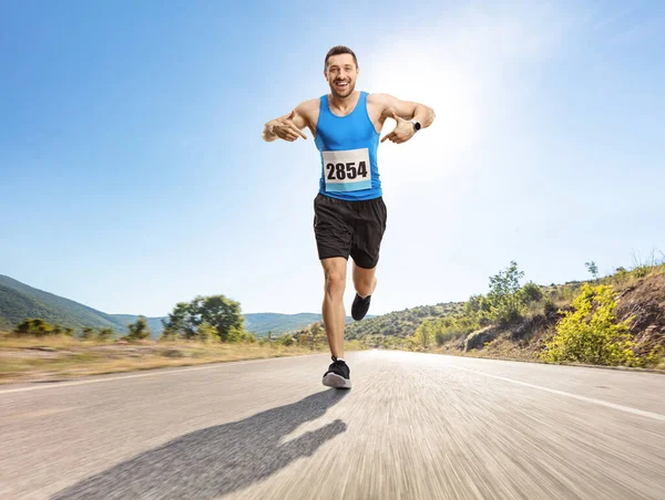 一个健康的年轻人在一条开阔的路上跑马拉松 指着他的号码 他的全长画像 — 图库照片