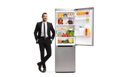Beyaz arka planda izole edilmiş açık bir buzdolabına yaslanmış takım elbiseli genç bir iş adamının tam boy portresi.