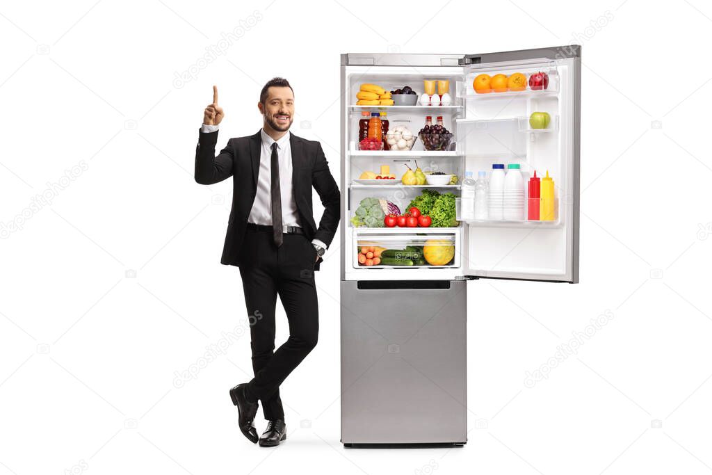 Full length portrait of a businessman in a suit leaning on an open fridge and pointing up isolated on white background