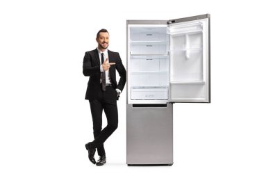 Takım elbiseli bir adamın uzun bir portresi boş bir buzdolabına dayanıyor ve beyaz arka planda izole edilmiş bir yeri işaret ediyor.