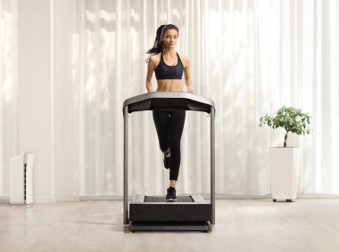 Bir odada koşu bandında egzersiz yapan zayıf bir genç kadının tam boy portresi. 