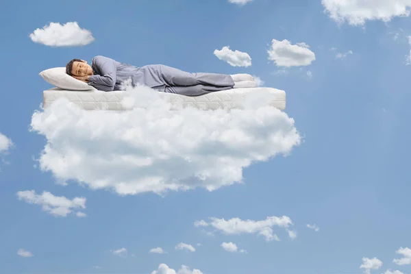Man in pajamas sleeping on a mattress and floating on clouds and a blue sky
