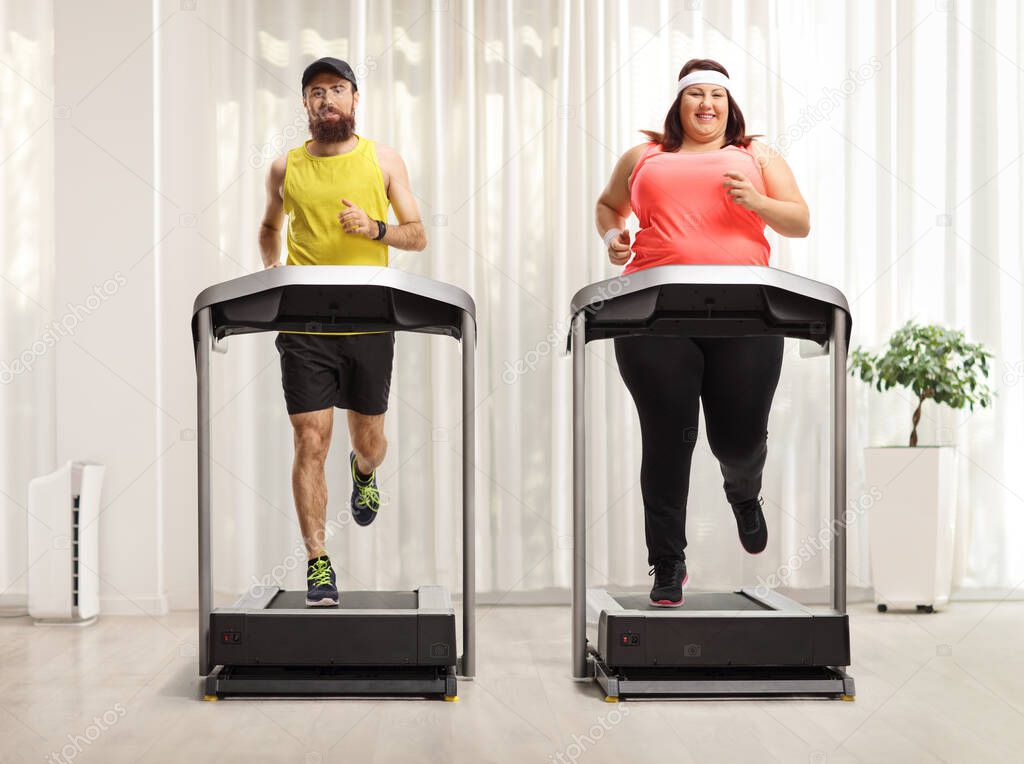 Full length portrait of a bearded man and a corpulent woman exercising on treadmills in a room 