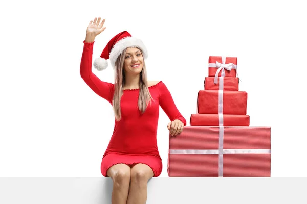 赤いドレスを着た魅力的な若い女性と 白い背景に孤立したプレゼントと手を振ってパネルに座っているサンタクラスの帽子 — ストック写真