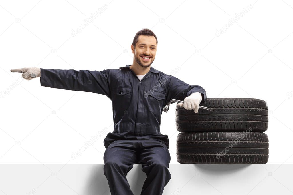 Auto mechanic worker sitting on a panel with tires and pointing to the side isolated on white background