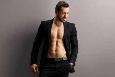 Siyah takım elbiseli genç adam gri bir duvara üstsüz poz veriyor.