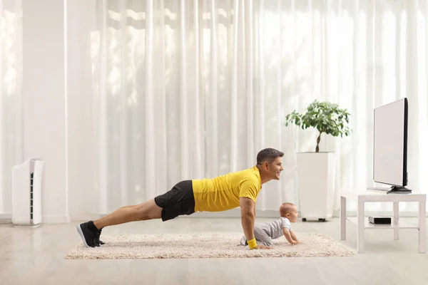 父亲在电视机前锻炼 婴儿在白色背景的地毯上爬行 — 图库照片