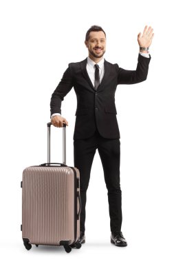 Full length portrait of a young professional man in suit and tie holding a suitcase and waving isolated on white background