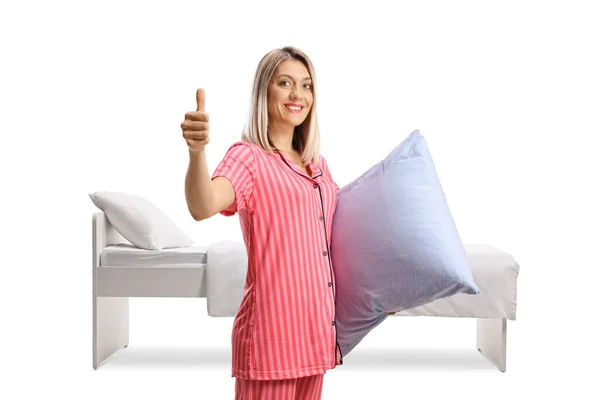 枕を持つベッドの前に立つパジャマ姿のブロンド女性と白い背景に孤立した親指を示す — ストック写真