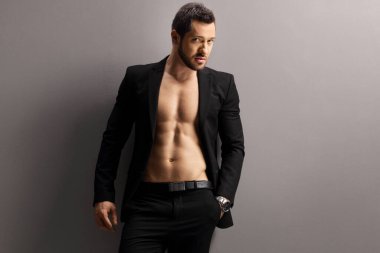Siyah takım elbiseli bir erkek model üstsüz poz veriyor ve gri bir duvara bakıyor.