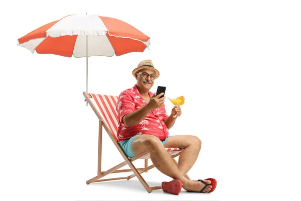 一个拿着鸡尾酒和手机的成熟男人坐在沙滩上的椅子上 被白色的背景遮掩着 — 图库照片