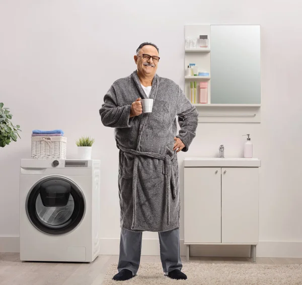 Mature man wearing a robe and standing inside a bathroom with a cup of coffee