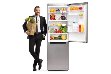 Beyaz arka planda izole edilmiş buzdolabına yaslanmış market poşeti taşıyan genç bir iş adamının tam boy portresi.