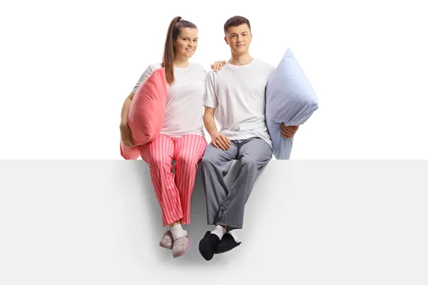 白い背景に隔離されたパネルに座って抱擁でポーズをとっているパジャマ姿の若いカップルの完全な長さの肖像画 — ストック写真