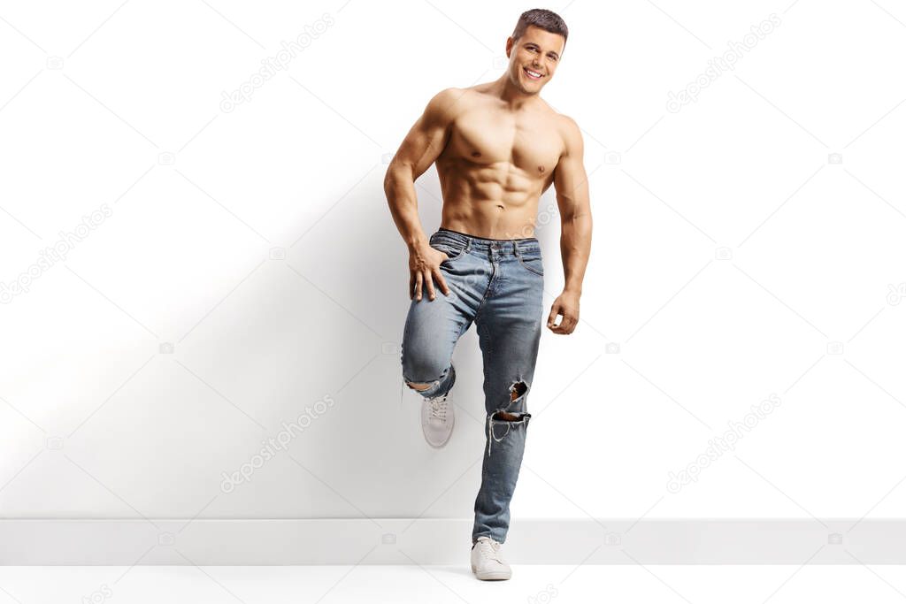 Full length portrait of a guy with muscular body posing shirtless and leaning on a wall isolated on white background