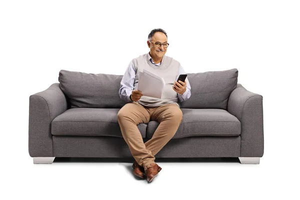 一个成熟的男人坐在沙发上 手里拿着手机 手里拿着用白色背景隔开的纸质文件 — 图库照片