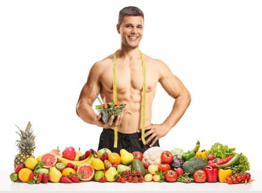 Bir yığın meyve ve sebzeli vücut geliştirici salata kasesi tutuyor ve beyaz arka planda izole edilmiş kameraya bakıyor.