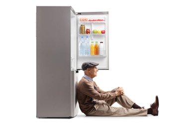 Yaşlı bir adam yerde oturuyor ve beyaz arka planda izole edilmiş bir buzdolabına yaslanıyor.