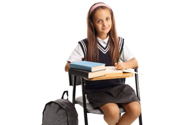 Okul üniformalı solak bir kız beyaz arka planda izole bir şekilde oturuyor ve okul ödevlerini yazıyor.